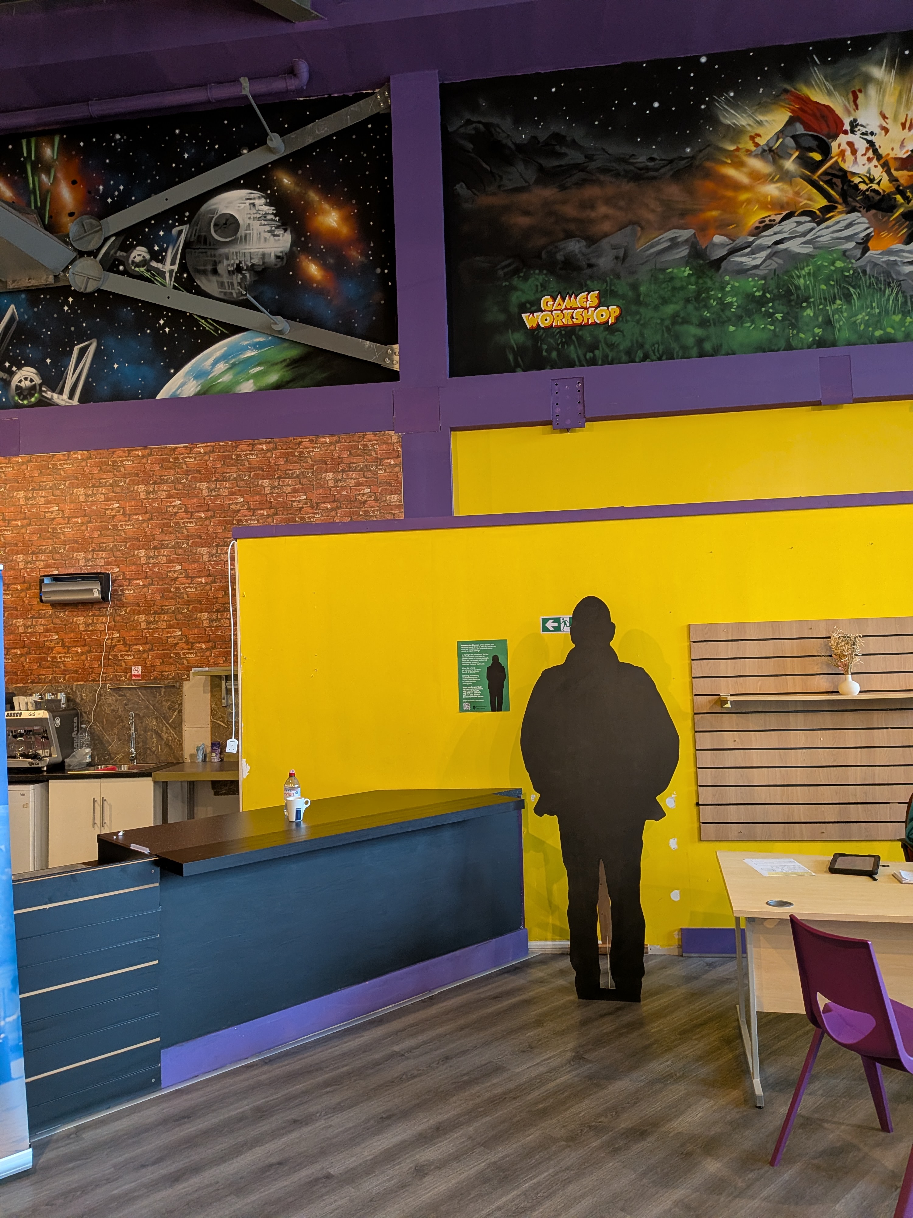 A black coloured shape of a man stands in a cafe with kitchen and chairs and tables around.