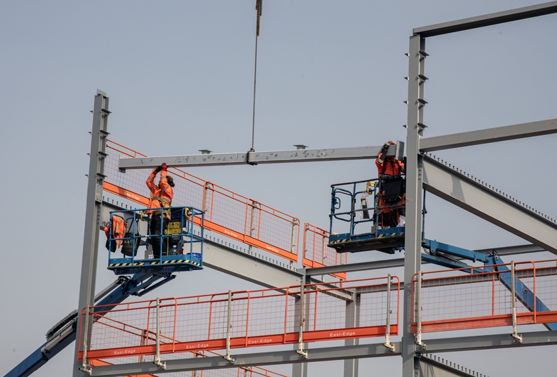 Two building site workers move a steel beam into place while it is being lifted by a crane. 