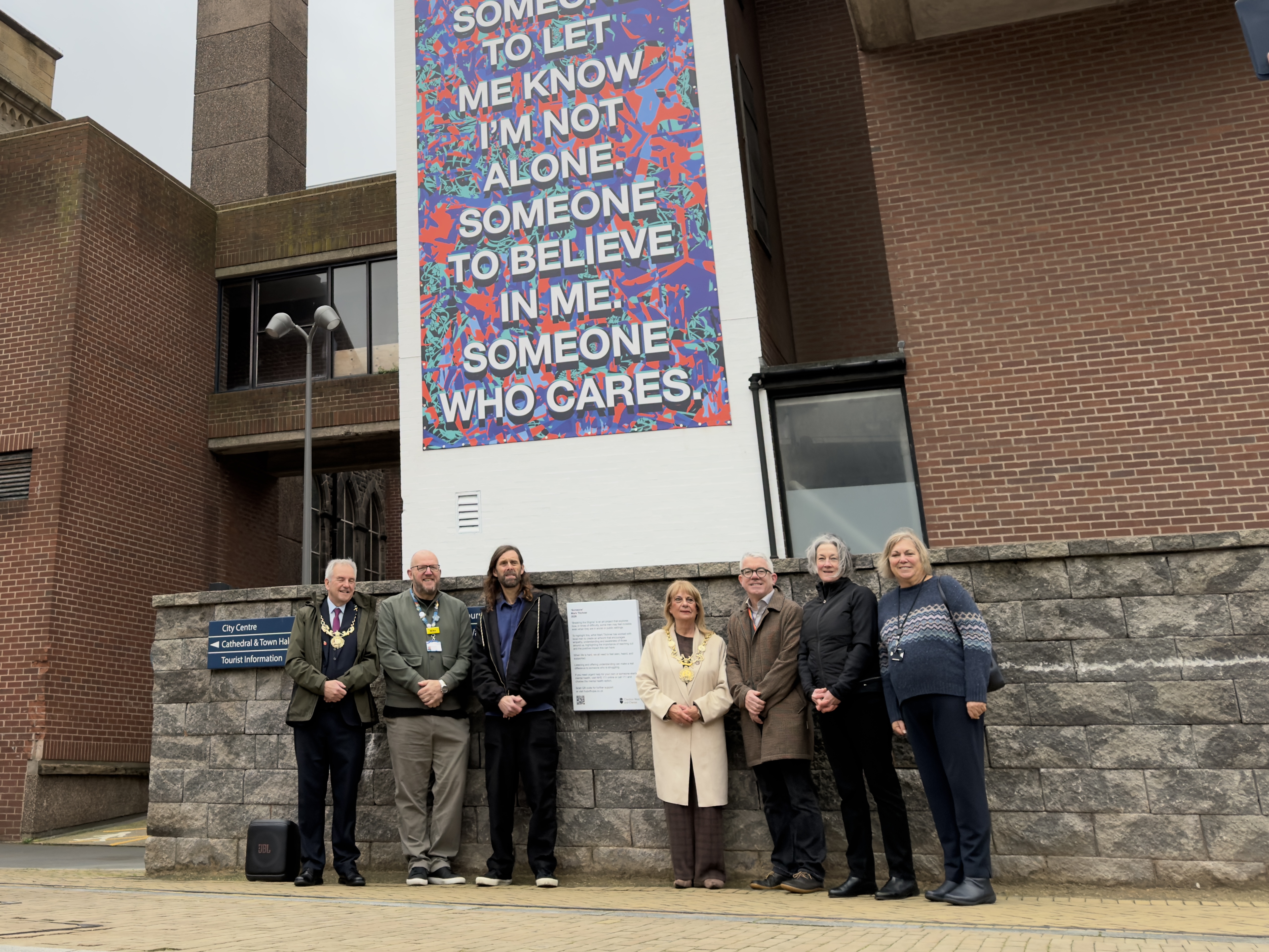 A picture of seven people standing outside a building with a mural in the background. The Mural has a quote on it.