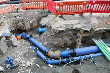Blue gas and water canalisation pipes showing on a hole in the ground and tarmac at a roadworks site.