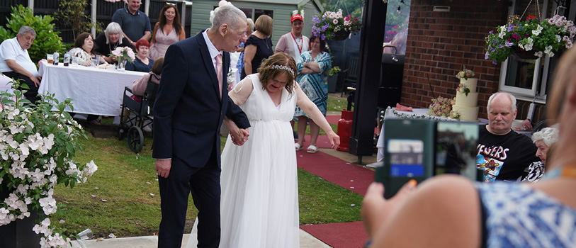 A man and woman holding hands. The man is wearing a suit and the woman is in a white long formal dress.