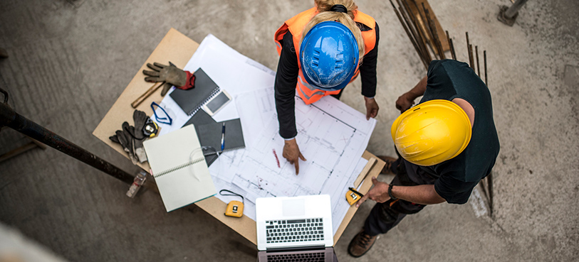 Two construction professionals in blue and yellow hard hats examining the blueprints on construction site.