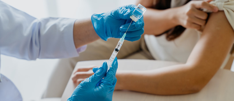 Person wearing blue gloves giving a COVID-19 vaccine. A person with their sleeve rolled up and arm on the table seen in the background.