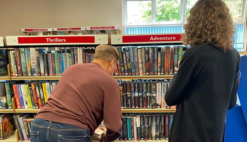 A man bending down to put books on a book shelf in a library. A woman is standing nearby watching.