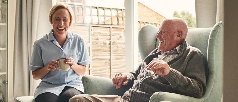 Indoor shot of smiling senior man and female carer enjoying coffee in living room.