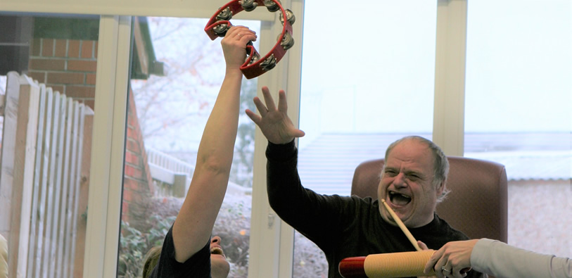 A man sat on a chair smiling, next to him a person is holding a tambourine in the air and the man's arm is raised trying to reach the tambourine.