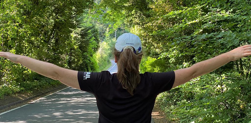 Woman in black T-shirt and white cap stands in a tree-lined country lane with arms outstretched
