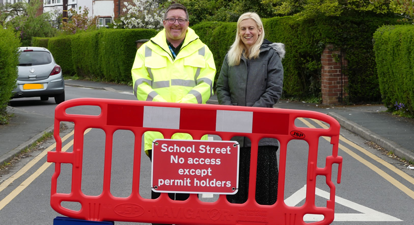 Two school staff members standing behind a red barrier, a sign on the barrier reads 'School Street, no access except permit holders'.