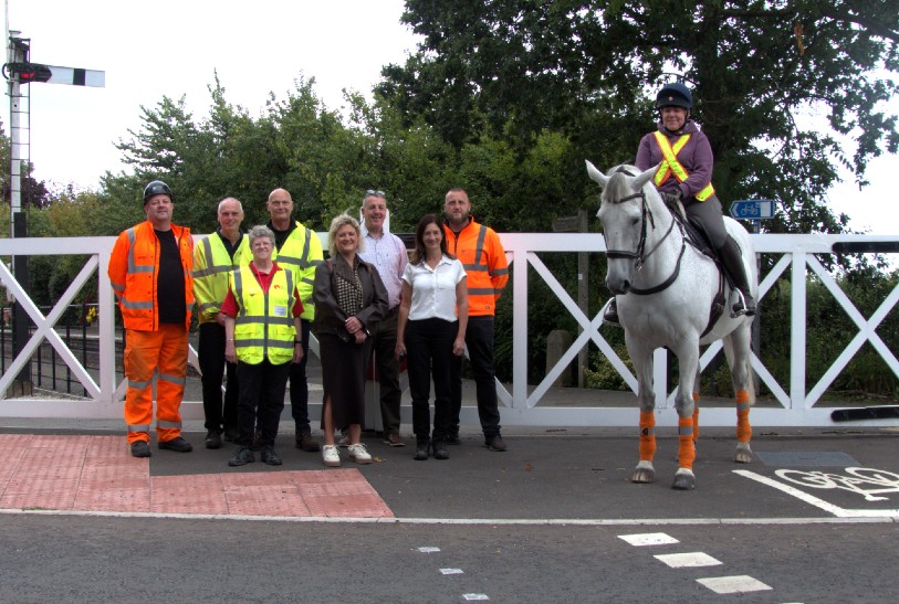 Group of people, some wearing hi-his jackets, standing in front of a white level crossing with a rider on a grey horse.