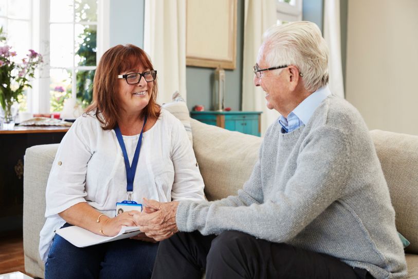 Caring lady in white top and wearing blue lanyard holds hand of elderly gentleman wearing a grey jumper and glasses