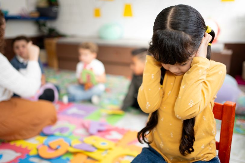 Dark haired little girl, sat on chair, hands over her ears, looking sad. Other children playing on floor with a teacher