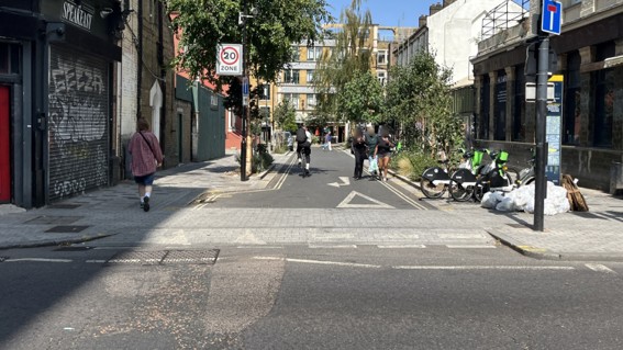 Retrofitted continuous crossing across a side street, with a level pedestrian route extending across the junction. Trees, cycle parking and pedestrians line the street, with vehicle access maintained at low speeds.