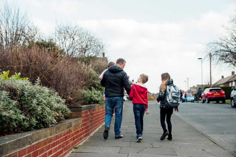 A man is walking up a road with two children by his side and he's carrying a smaller child. 