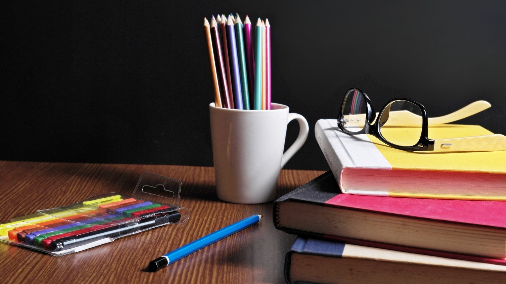 pencils a pile of books and a pair of glasses on a wooden desk